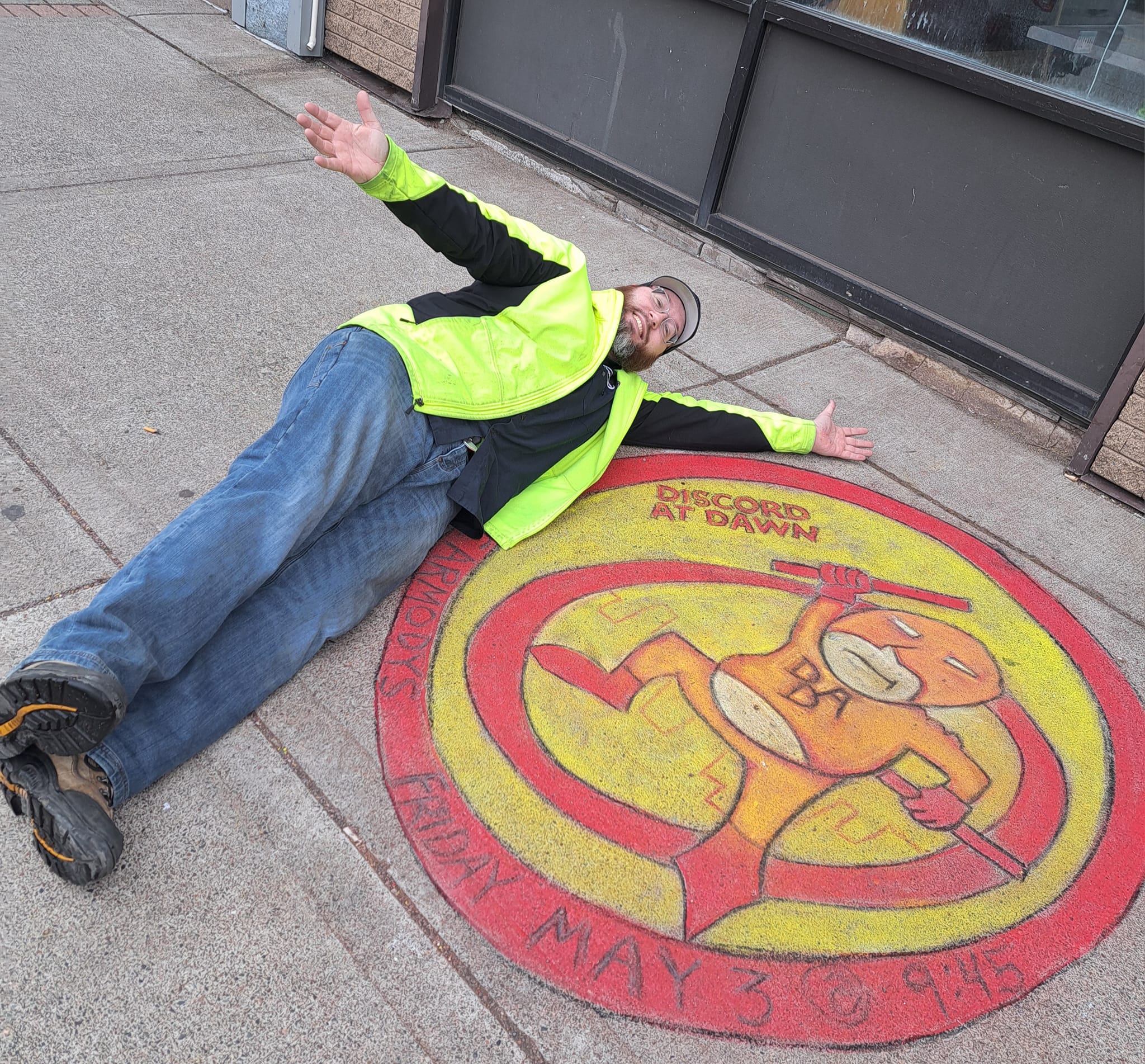 Joel laying on the sidewalk next to chalk art of Discord at Dawn's logo advertising our homegrown show in 2024.