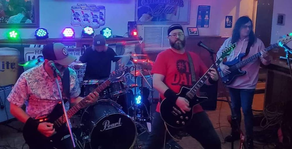 The band playing live in a smoky bar.  From left to right: Andy in a white hawaiian shirt with palm trees, David in a black t-shirt, Joel in a red t-shirt that says #1 DAD, and Robin in a pink sweatshirt.