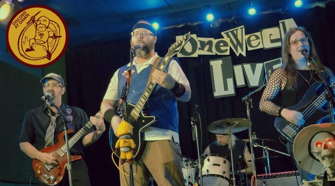 The band playing live. From left to right: Andy in black playing a red guitar, Joel with a headband and a vest playing a black guitar, David sitting at the drums in a black tank top, and Robin in a black tank top with fishnet arms playing a blue bass guitar.